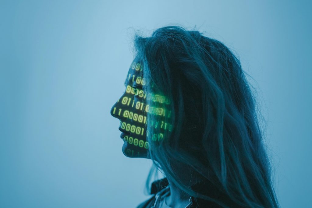 Photo by cottonbro studio on Pexels. Shows a woman white pale hair with 1s and 0s obscuring her face. The projected 1s and 0s are gree, while the rest of the image is shades of blue.