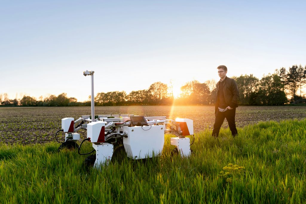 Photo by ThisisEngineering on Unsplash. A man in a field in the evening walking behind a robot.